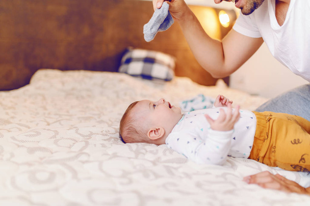 Boldog jóképű kaukázusi fiatal apa kezében apró baba zokni és készül átöltözni a nevető aranyos 6 hónapos fia. Hálószoba belső. - Fotó, kép