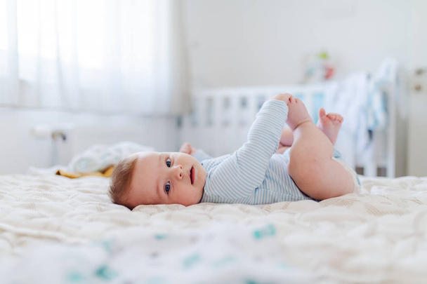 Side view of adorable caucasian 6 months old baby boy dressed in bodysuit holding his feet and looking at camera while lying on bed in bedroom. - Photo, Image