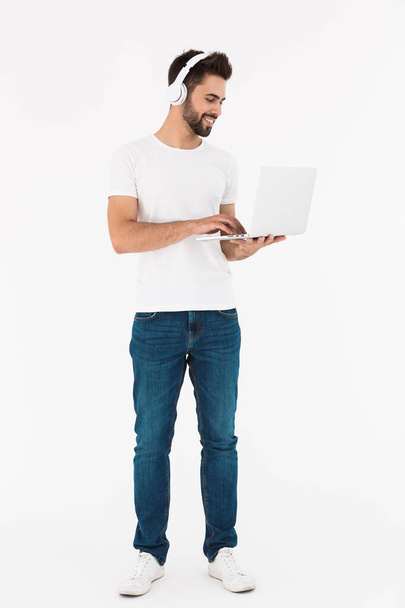 Full length image of pleased satisfied man smiling while using laptop and headphones - Foto, Imagem