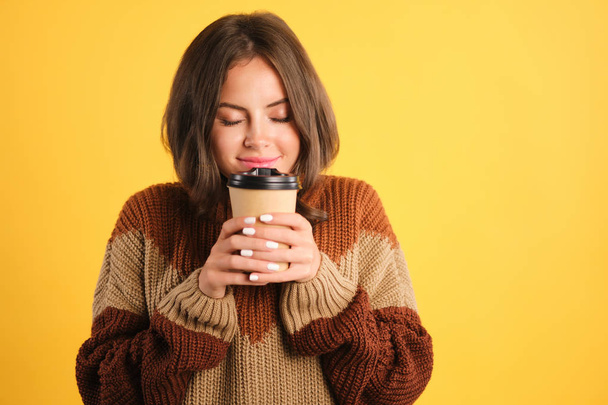 Привлекательная улыбающаяся девушка в уютном свитере мечтательно нюхает кофе на желтом фоне
 - Фото, изображение