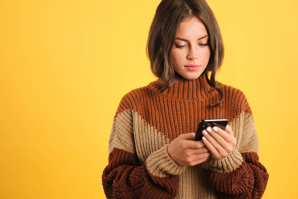 Kaunis tyttö viihtyisä villapaita luottavaisin mielin käyttämällä matkapuhelinta keltaisella taustalla
 - Valokuva, kuva