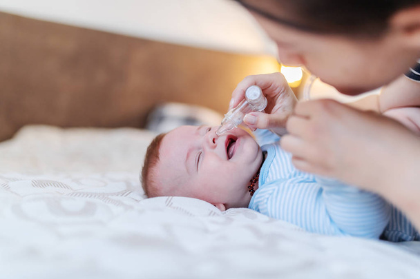 Primer plano de cuidar orgullosa madre caucásica usando la bomba nasal para limpiar la nariz de su bebé. Bebé acostado en la cama vestido con traje de cuerpo
. - Foto, Imagen