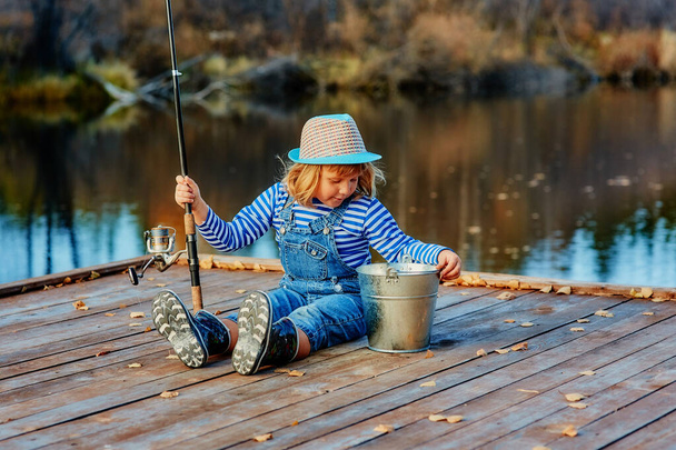 A little girl with a fishing rod looks at the catch of fish in a bucket - Zdjęcie, obraz