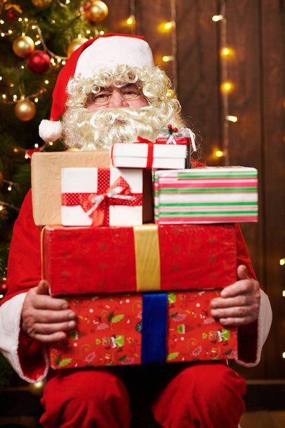 Père Noël posant avec de nombreux cadeaux, assis à l'intérieur près de l'arbre de Noël décoré avec des lumières Joyeux Noël et Joyeuses Fêtes
! - Photo, image