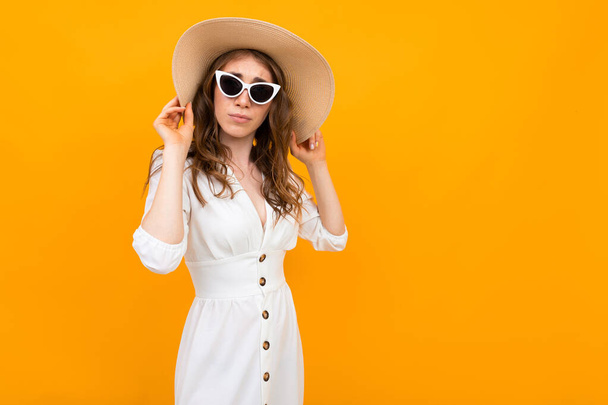 mujer joven de moda posando en estudio contra naranja usando vestido blanco, sombrero de paja y gafas de sol
 - Foto, Imagen