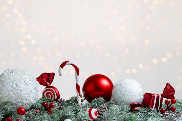 Composição branca e vermelha de Natal com globos, doces, cana-de-açúcar e pirulito sobre alguns abetos nevados ou ramos de pinho com cones. luz bokeh fundo e espaço de cópia
. - Foto, Imagem