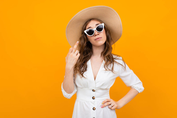 fashionable young woman posing in studio against orange wearing white dress, straw hat and sunglasses - Foto, afbeelding