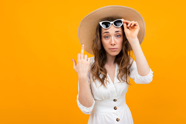 Stüdyoda beyaz elbise, hasır şapka ve güneş gözlüğü takmış, turuncuya karşı poz veren şık bir genç kadın. - Fotoğraf, Görsel