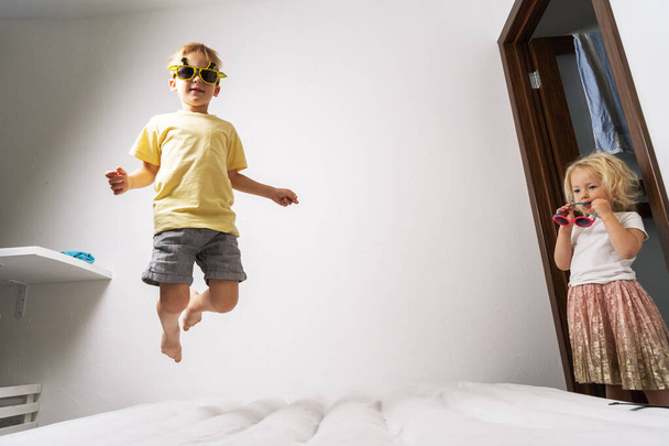 A little boy in yellow sunglasses and a yellow skirt jumps on a white mattress. Studio portrait on a white background. - Фото, изображение