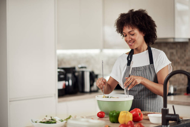Attractive mixed race woman in apron and with curly hair mixing vegetables in bowl while standing in kitchen at home. - Photo, image