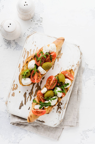 Caprese bruschetta toasts with cherry tomatoes, mozzarella, olives and basil on white plate old light background. Top view. - Foto, Imagem