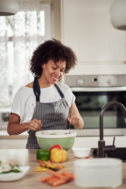 Attractive mixed race woman in apron and with curly hair mixing vegetables in bowl while standing in kitchen at home. - Foto, Imagem