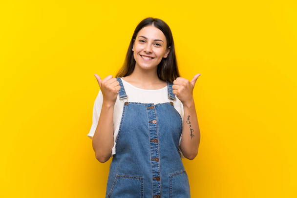 jovem mulher no dungarees sobre isolado amarelo fundo com polegares para cima gesto e sorrindo
 - Foto, Imagem