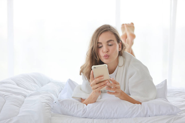 Νεαρή ελκυστική καυκάσια γυναίκα φορώντας λευκό πιτζάμα χρησιμοποιώντας το κινητό τηλέφωνο και δίνοντας φιλί στο κρεβάτι στο υπνοδωμάτιο (χαλάρωση με την έννοια της τεχνολογίας) - Φωτογραφία, εικόνα