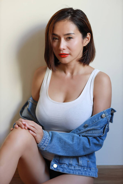 vista lateral joven adulto asiático mujer, labio rojo y pelo corto en vestido blanco y jean azul con cara de belleza y mirando cámara con pared blanca - Foto, Imagen