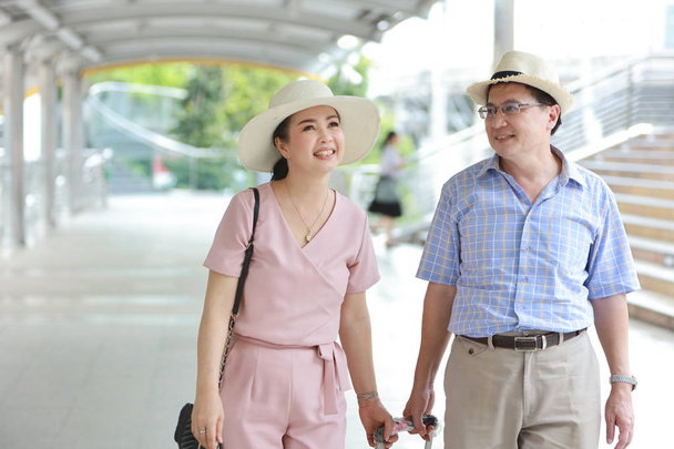 asiatisches Reisepaar Mann und Frau in rosa und blauem Hemd mit weißem Hut und Gepäck spaziert im Rentenalter glücklich lächelnd durch die Innenstadt - Foto, Bild