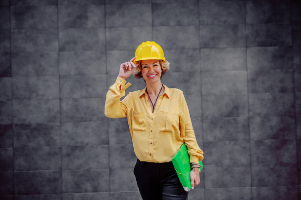 Чудова біла усміхнена старша жінка архітекторка з шоломом на голові і документами під пахвою позує перед сірою стіною на відкритому повітрі
. - Фото, зображення