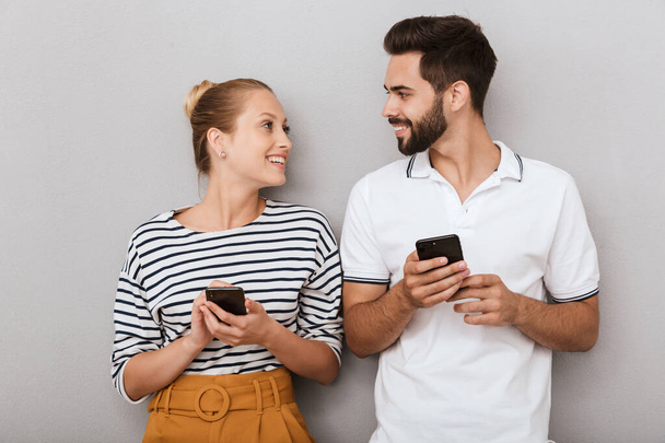 Pleased happy young positive loving couple friends man and woman posing indoors isolated over grey background using mobile phones. - Foto, Imagem