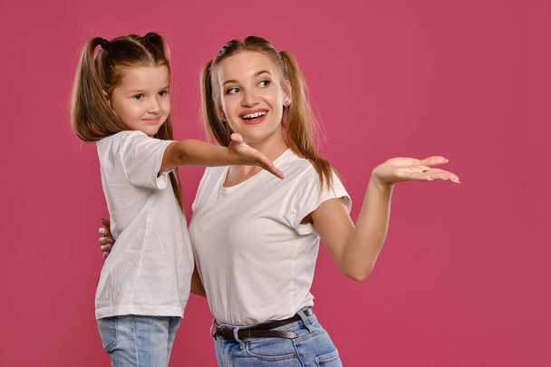白いTシャツと青いデニムジーンズを着た面白いポニーテールの母と娘はピンクのスタジオの背景にポーズをとっています。クローズアップショット. - 写真・画像