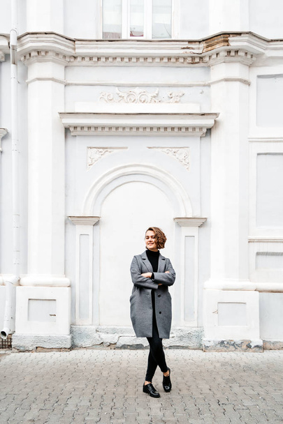 girl in gray coat and black jeans stands near building - Zdjęcie, obraz