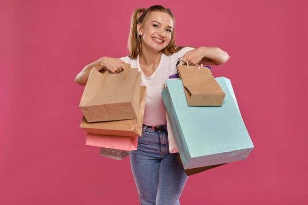 Blond vrouwtje met paardenstaarten, gekleed in wit t-shirt en jeans poserend tegen een roze achtergrond met pakjes. Een close-up. Oprechte emoties, winkelen. - Foto, afbeelding