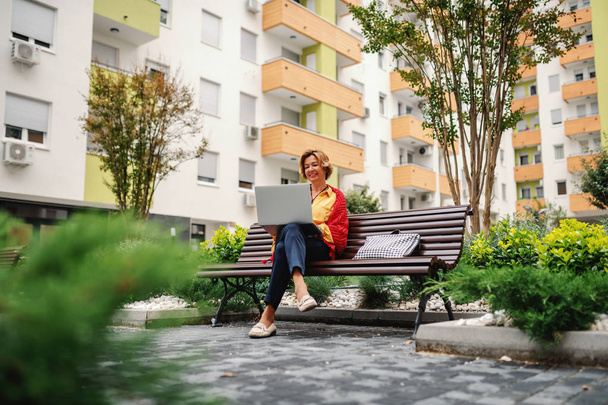 Beautiful smiling caucasian senior woman sitting on bench in park and using laptop. - Foto, Imagem