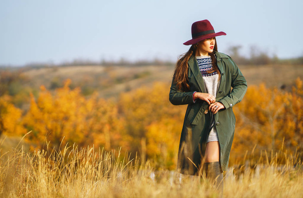 Piękna młoda, stylowa dziewczyna w płaszczu spaceruje jesienią w parku. Dziewczyna jest ubrana w zielony płaszcz i czerwony kapelusz. Piękny wieczór. Jesienna moda. Styl życia. Wysoki portret mody. - Zdjęcie, obraz