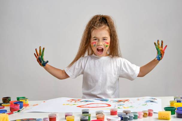 Little girl in white t-shirt sitting at table with whatman and paints on it, posing with painted face and hands. Isolated on white. Medium close-up. - Photo, Image