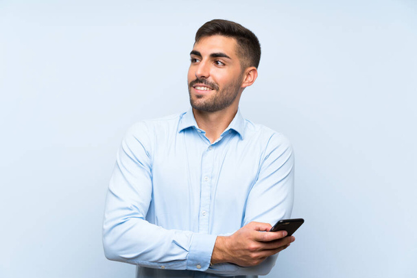 Молодой красивый мужчина с мобильным телефоном над изолированной голубой стеной смотрит вверх, улыбаясь
 - Фото, изображение
