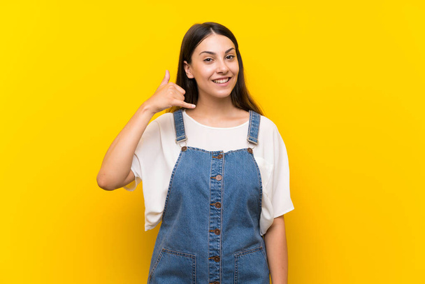 junge Frau in Latzhose über isoliertem gelben Hintergrund mit Handygesten. rufen Sie mich zurück - Foto, Bild
