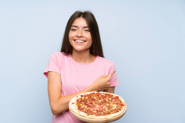 Csinos fiatal lány kezében egy pizza felett elszigetelt kék fal felé mutató oldalán, hogy bemutassa a termék - Fotó, kép
