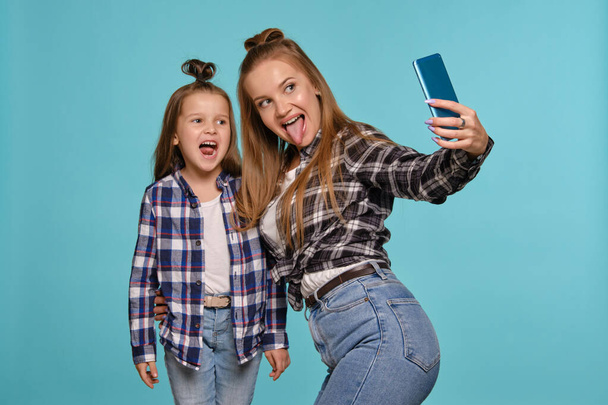 Mom and daughter dressed in checkered shirts and blue denim jeans are making selfie while posing against a blue studio background. Close-up shot. - Photo, Image