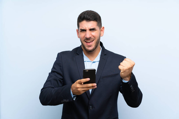 schöner Mann über isoliertem blauem Hintergrund mit Telefon in Siegesposition - Foto, Bild
