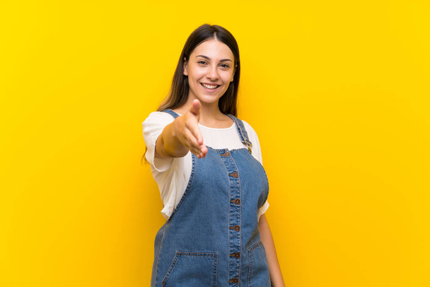 junge Frau in Latzhose vor isoliertem gelben Hintergrund schüttelt Hände, um ein gutes Geschäft abzuschließen - Foto, Bild