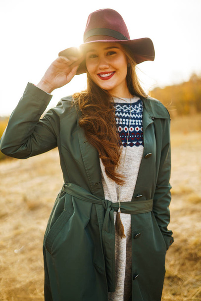 Paltolu güzel, genç, şık bir kız sonbaharda parkta yürüyor. Kız yeşil bir ceket ve kırmızı bir şapka giyiyor. Güzel bir akşam. Sonbahar modası. Yaşam tarzı. Yüksek moda portresi. - Fotoğraf, Görsel
