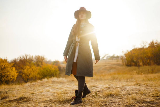 Beautiful young stylish girl in a coat walks in the autumn in the park. The girl is dressed in a green coat and a red hat. Beautiful evening. Autumn fashion. Lifestyle. High fashion portrait. - Foto, Bild