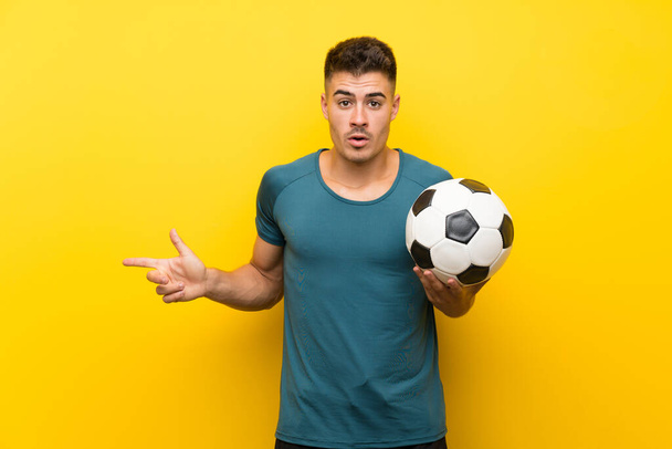 Beau jeune joueur de football homme sur fond jaune isolé surpris et pointant côté
 - Photo, image