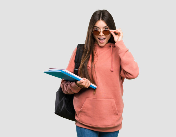 メガネをかけたノートを持つ若い学生の女性と孤立した灰色の背景に驚いた - 写真・画像