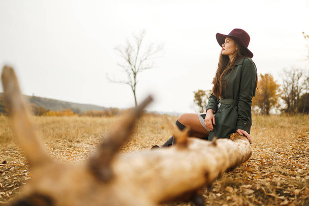 Beautiful young stylish girl in a coat walks in the autumn in the park. The girl is dressed in a green coat and a red hat. Beautiful evening. Autumn fashion. Lifestyle. High fashion portrait. - Photo, Image