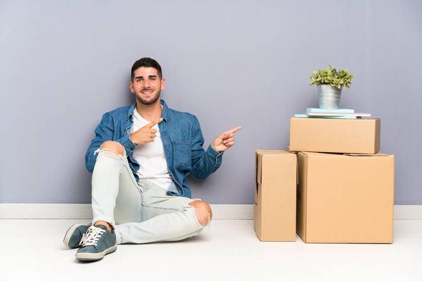Handsome young man moving in new home among boxes pointing finger to the side - Фото, зображення