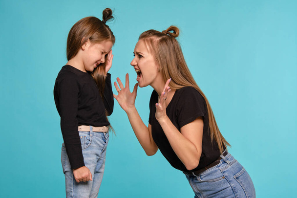 Mom and daughter with a funny hairstyles, dressed in black shirts and blue denim jeans are posing against a blue studio background. Close-up shot. - Fotó, kép