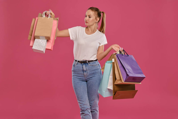 Blonde woman with ponytails, dressed in white t-shirt and jeans posing against a pink background with packages. Close-up. Sincere emotions, shopping. - Zdjęcie, obraz
