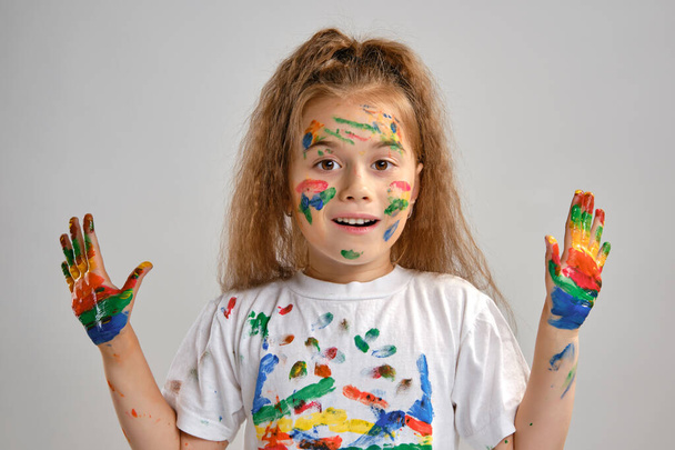 Little girl in white t-shirt is posing standing isolated on white and gesticulating with her painted in different colors palms. Art studio. Close-up. - Photo, image