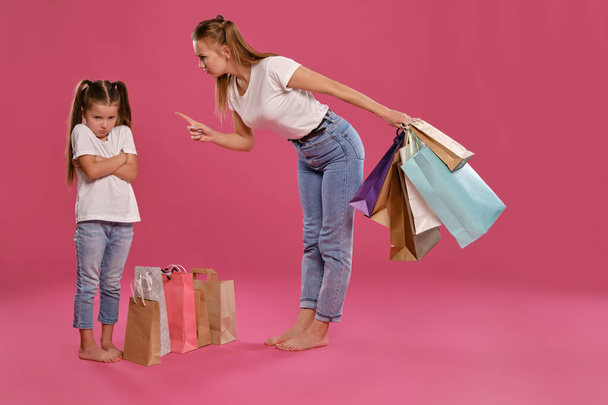 Mom and daughter with ponytails, dressed in white t-shirts and blue jeans are posing against pink background with packages in hands. Full length. - Foto, Imagem