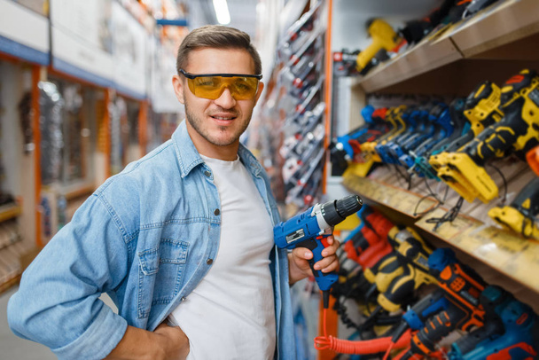 Mężczyzna kupujący elektryczny śrubokręt w sklepie z narzędziami. Klienci patrzą na towary w sklepie diy, zakupy w budynku supermarketu - Zdjęcie, obraz