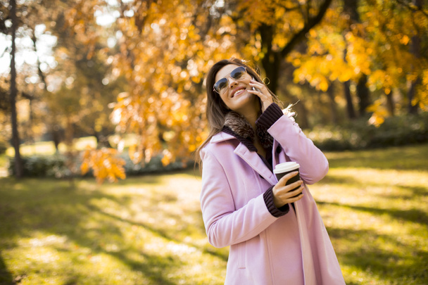 Pembe ceketli neşeli genç bir kadın güneşli parkta telefonunu kullanıyor ve kahve içiyor. - Fotoğraf, Görsel