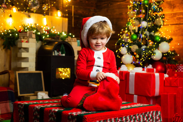Υπέροχο μωρό απόλαυσε τα Χριστούγεννα. Παιδί σε χριστουγεννιάτικο δέντρο και τζάκι παραμονή Χριστουγέννων. Οικογένεια με παιδιά που γιορτάζουν τα Χριστούγεννα στο σπίτι. Μικρό αγόρι στολίζει χριστουγεννιάτικο δέντρο και ανοίγει δώρα. - Φωτογραφία, εικόνα