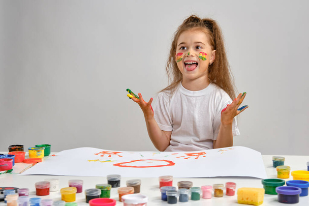 Little girl in white t-shirt sitting at table with whatman and colorful paints on it, showing her pained hands, tongue. Isolated on white. Close-up. - Foto, Imagen