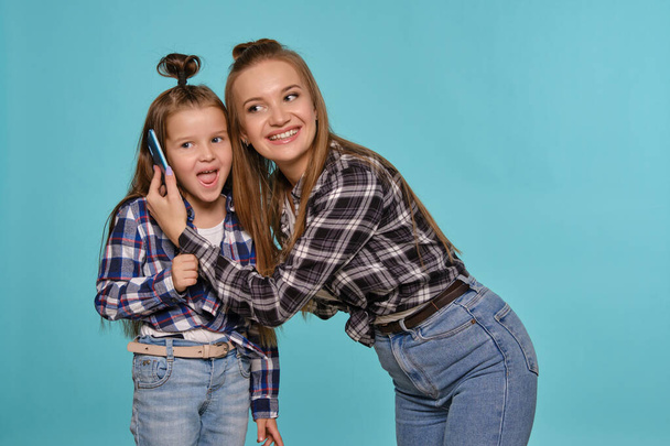 Mãe e filha vestidas com camisas de xadrez e jeans jeans jeans azuis estão usando smartphone enquanto posa contra um fundo estúdio azul. Close-up shot. - Foto, Imagem