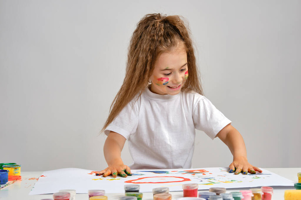 Petite fille en t-shirt blanc assise à table avec whatman et peint dessus, posant avec visage et mains peints. Isolé sur blanc. Gros plan moyen. - Photo, image